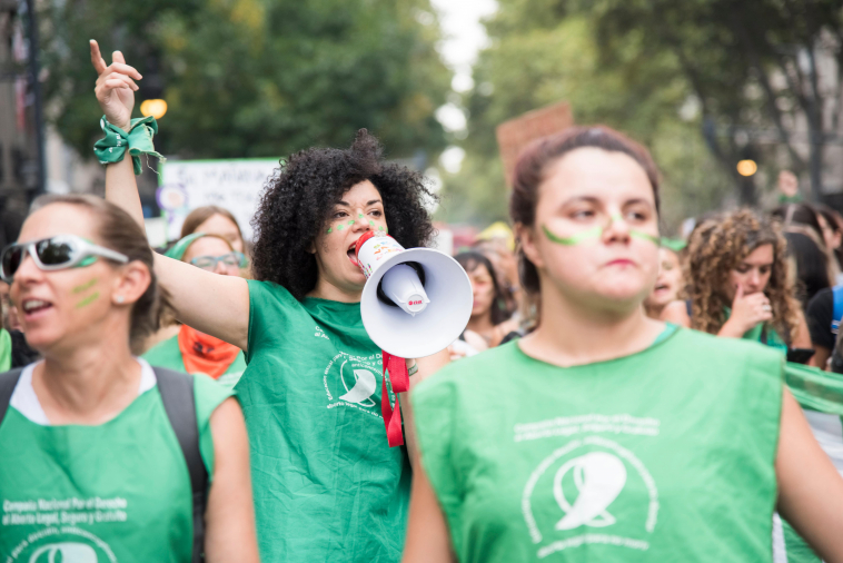 A young black woman speaking into a megaphone with a fist in the air. She is wearing a green top used as the symbol of the pro-choice movement in Latin America.  